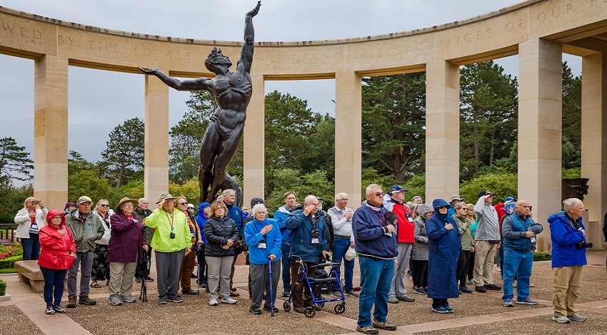 The group stands for a ceremony at the American Cemetery, with a statue in the background